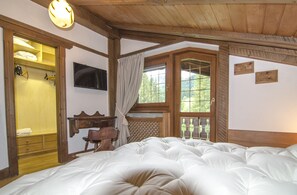 A bedroom with a large bed, wooden headboard, a wooden wardrobe, a desk with a chair, a window with a view of greenery, and a wall-mounted TV.