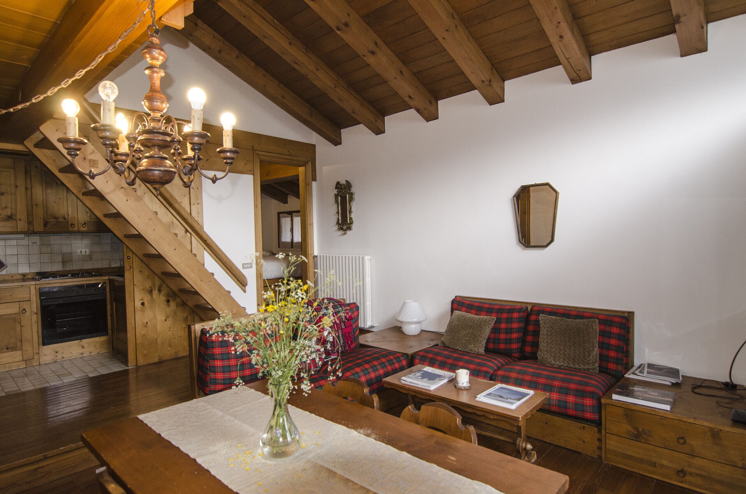 Ein gemütliches Zimmer mit einer Holztreppe, einer Couch mit karierten Kissen, einem Esstisch mit Blumenvase und einem Kronleuchter.