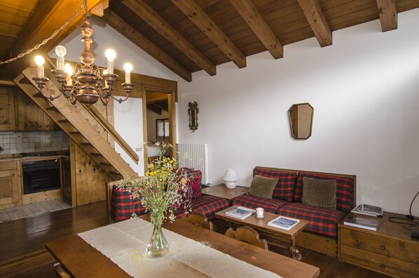 A cozy room with a wooden staircase, a sofa with plaid cushions, a dining table with a vase of flowers, and a chandelier.