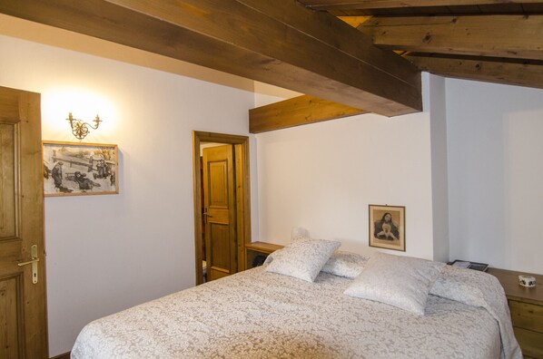 A bedroom with a bed, wooden ceiling beams, a door, and two framed pictures on the wall.