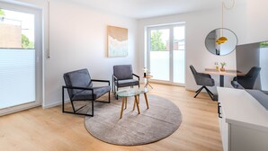 A modern living room with a glass table, two chairs, a round rug, a painting on the wall, and a round mirror.