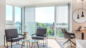 A modern living room with two grey armchairs, a glass coffee table, and a view of a cityscape through large windows. - Eversten- Penthouse (Oldenburg)