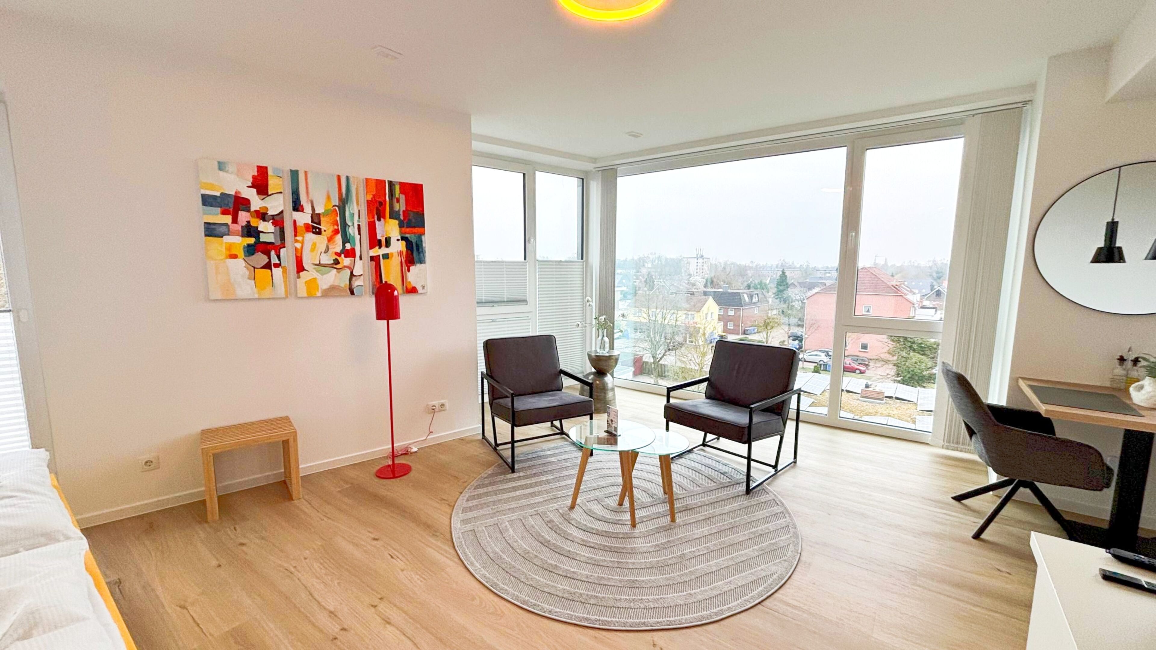 Un apartamento moderno con un ventanal grande, dos sillones, una mesita de centro y una lámpara roja de pie.