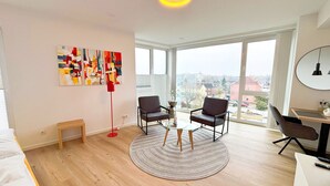 A modern apartment with a large window, two armchairs, a small coffee table, and a red floor lamp. - Eversten- Penthouse (Oldenburg)
