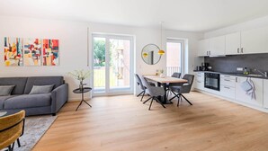 A modern apartment with a grey sofa, a dining area with a round table and chairs, a kitchen with white cabinets and a black countertop, and a large window with a view of the outside. - Kreyenbrück (Oldenburg)
