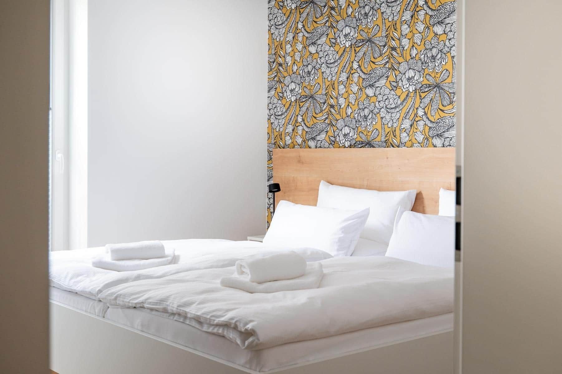 Una cama bien hecha con sábanas y toallas blancas, un cabecero de madera y un papel tapiz estampado con motivos florales.