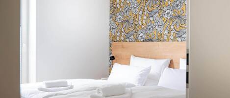 Una cama bien hecha con sábanas y toallas blancas, un cabecero de madera y un papel tapiz estampado con motivos florales.