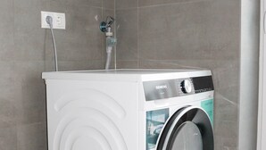 A compact, white front-loading washing machine in a corner of a room with grey tiled walls and floor. - Kreyenbrück (Oldenburg)