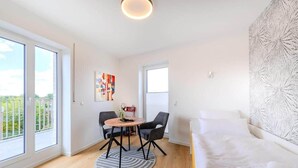 A modern, well-lit room with a large window, a round dining table with chairs, a bed with white bedding, and a patterned wall. - Bümmerstede (Oldenburg)