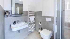 Un baño moderno con lavamanos, espejo y retrete blancos, y una cabina de ducha con mampara de vidrio.