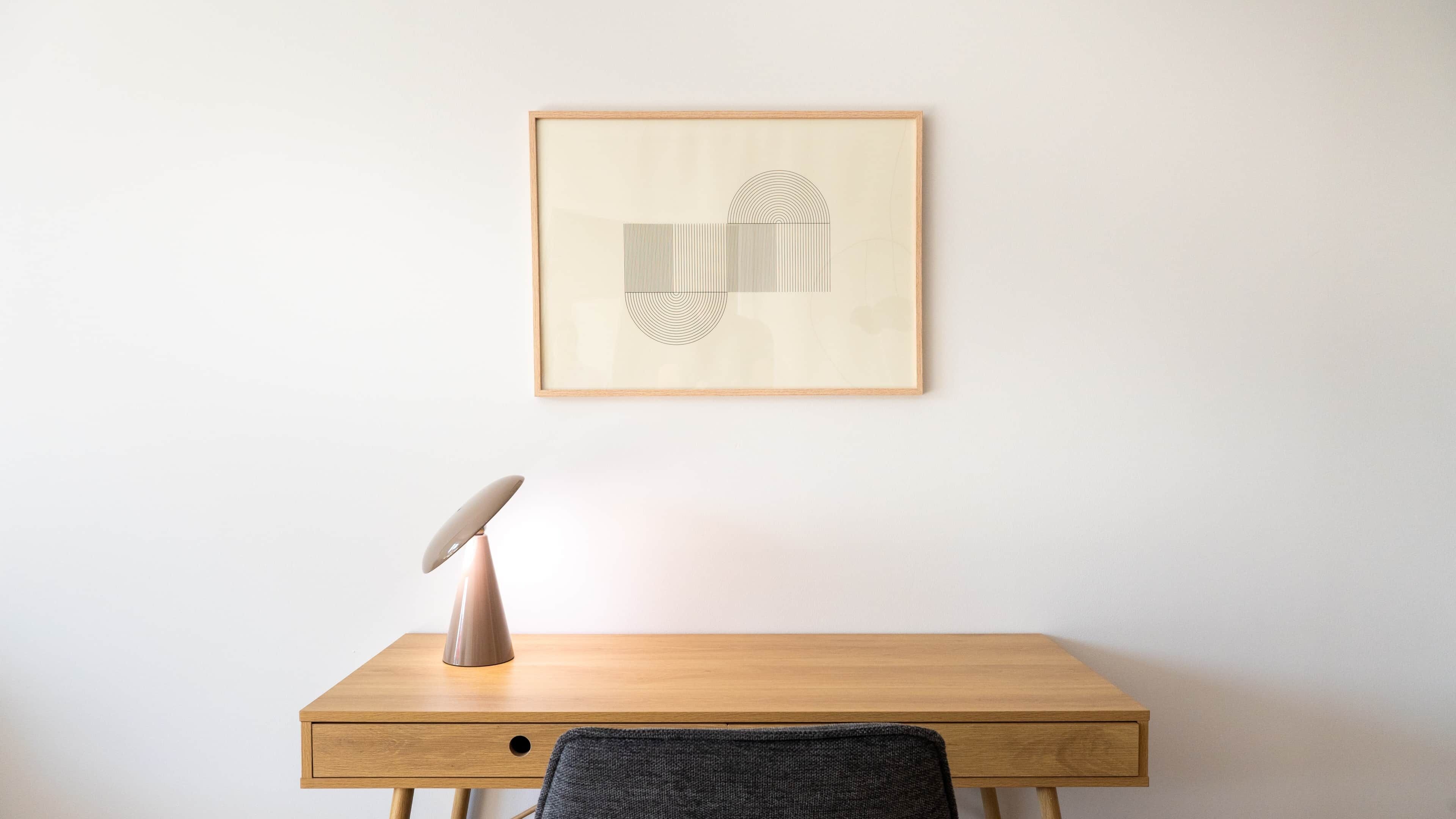 A wooden desk with a lamp and a framed abstract artwork on the wall.