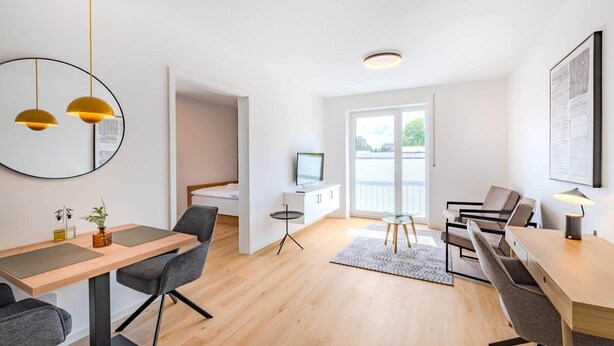 A modern, well-lit room with a dining area, a small seating area, and a bed in the background.