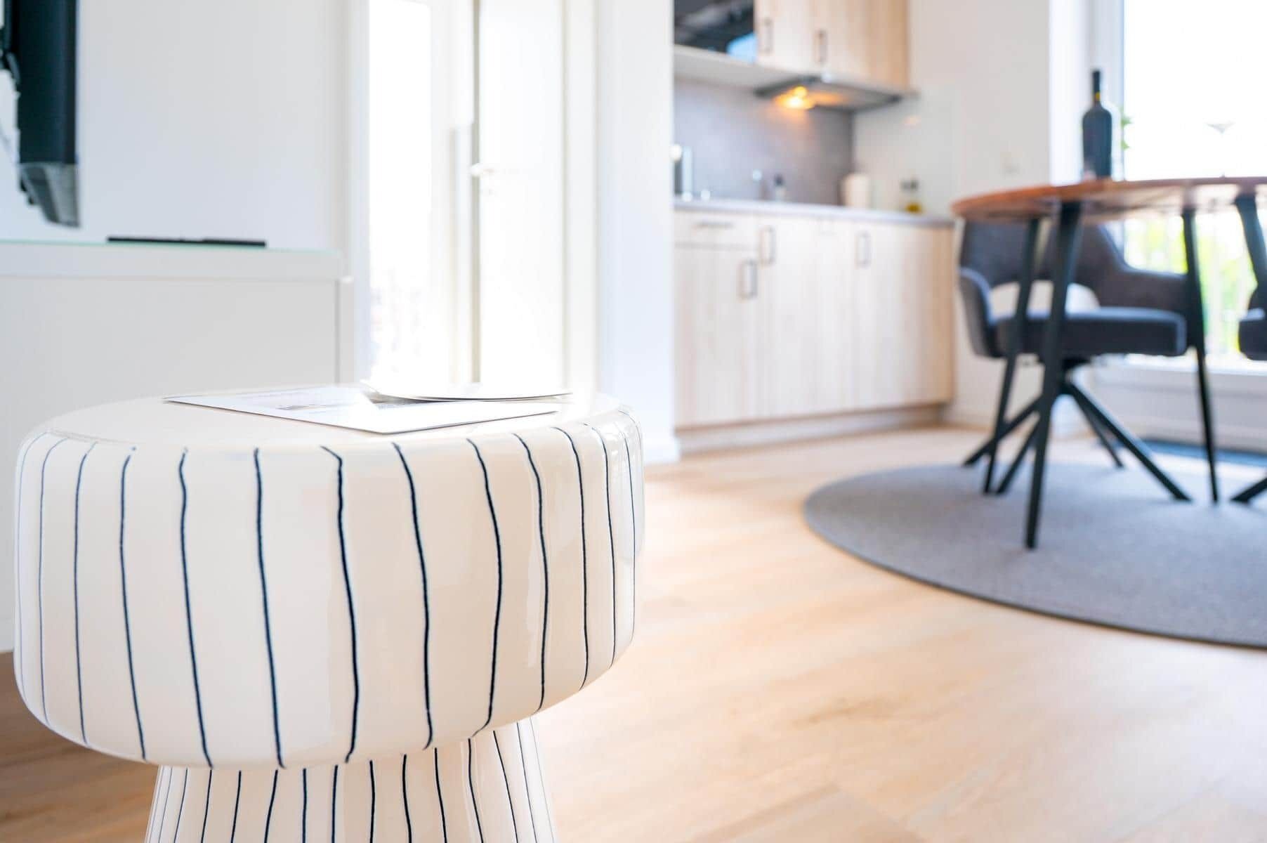 A modern kitchen with wooden cabinets, a dining area with a round table and chairs, and a white-striped ottoman with a laptop on it.