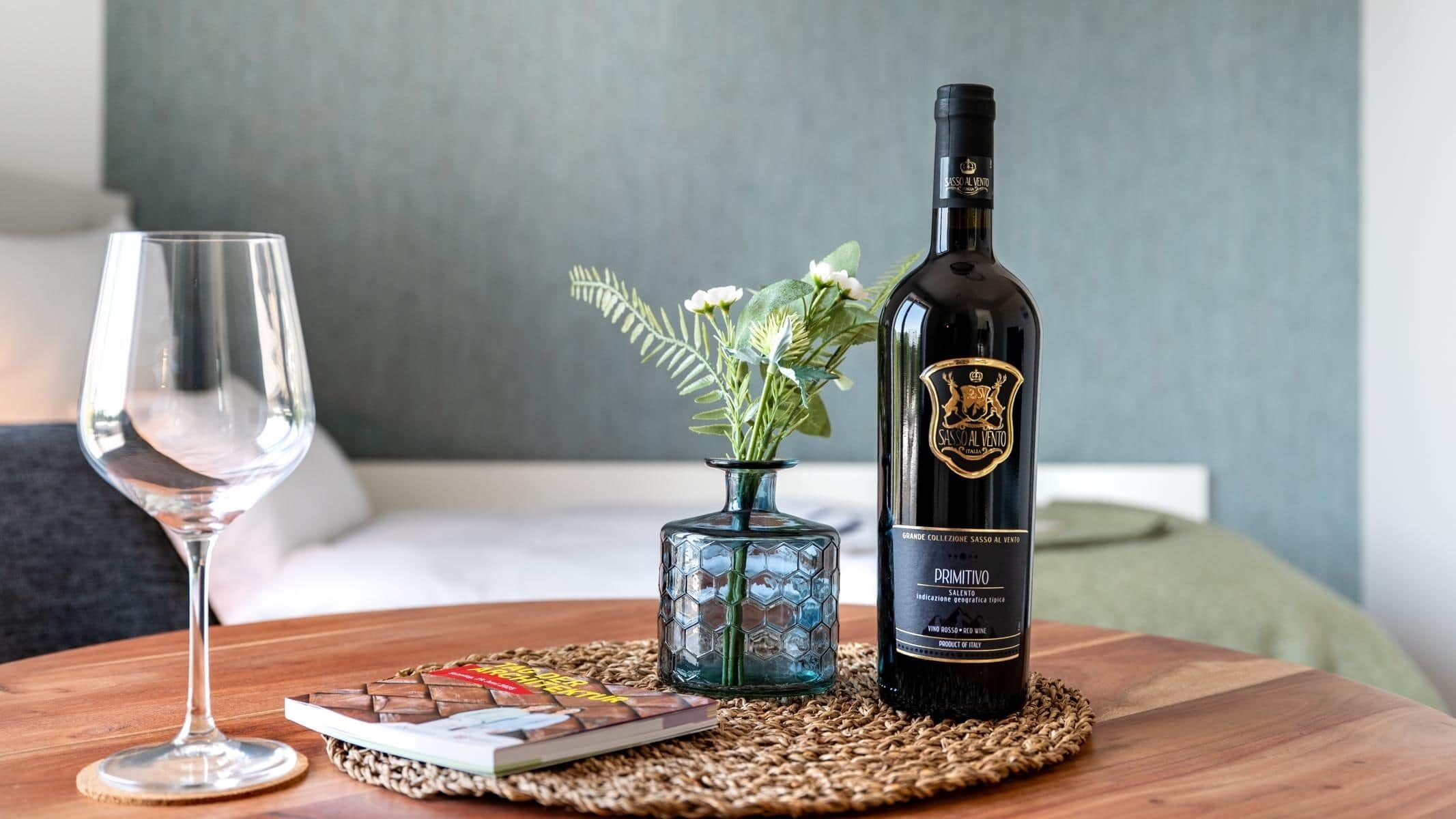 A bottle of wine, a glass, and a small vase with flowers on a wooden table.