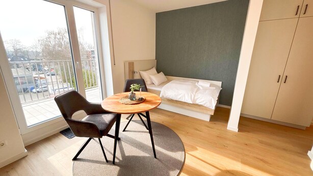 Une chambre moderne avec un lit, une petite table ronde ornée d’une plante, une chaise et une vue sur l’extérieur grâce à une grande fenêtre.