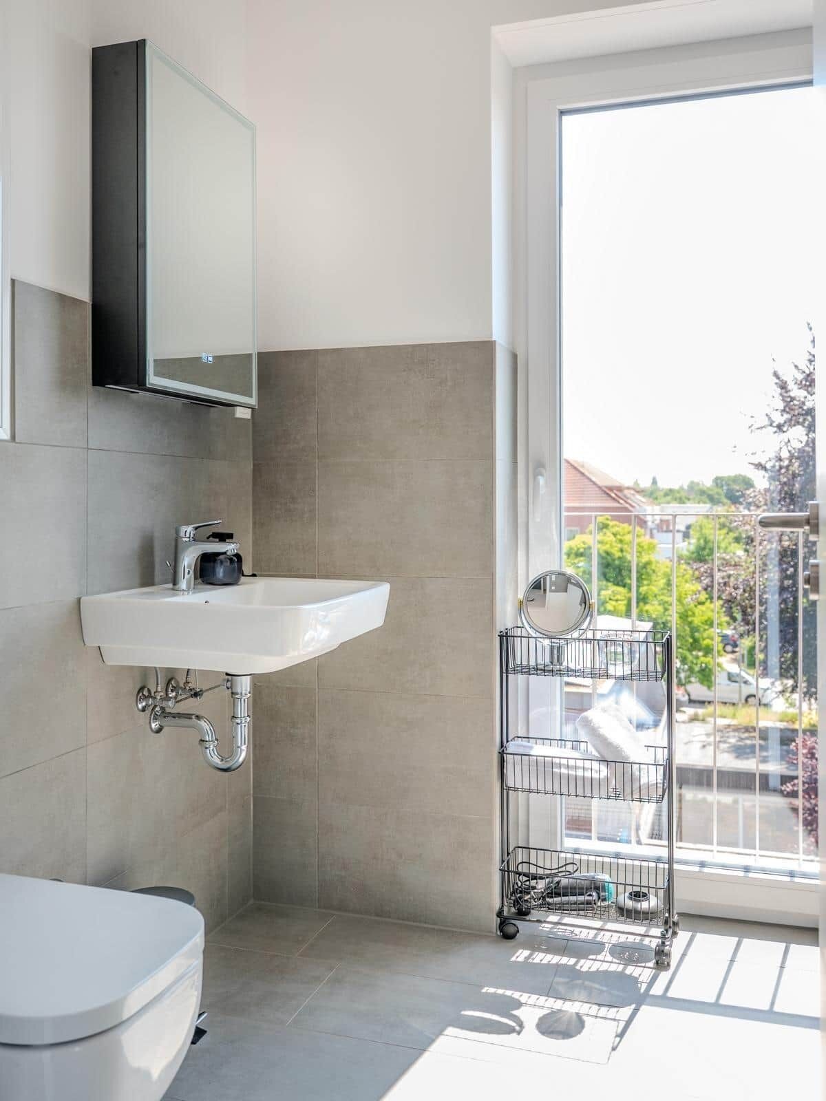 Un baño moderno con un lavamanos blanco, un inodoro y un espejo grande. Se aprecia una ventana con vistas a una zona residencial.