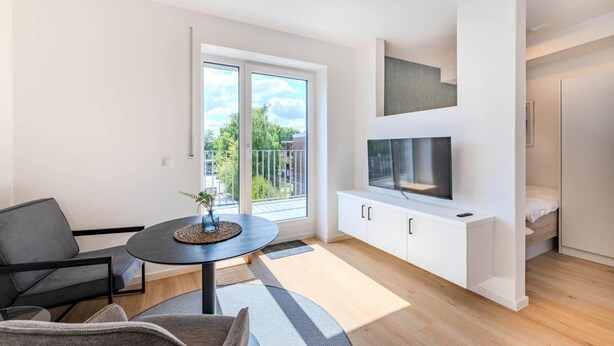 A modern apartment with a balcony, a TV mounted on the wall, and a dining area with a table and chairs.