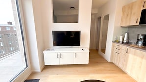 A modern kitchen with white cabinetry, a flat-screen TV mounted above the countertop, and a view of the city through a large window.