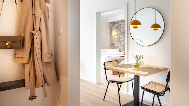 Habitación de hotel moderna con una mesa de madera, dos sillas negras, un espejo redondo con luces colgantes y una cama frente a una pared con un diseño.