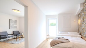 Un dormitorio moderno con una cama grande, una mesita de noche pequeña, dos sillas y un ventanal que deja entrar luz natural.