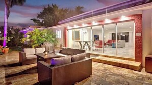 Un espacio exterior moderno con mobiliario de mimbre, un comedor con paredes de vidrio y vistas al interior.
