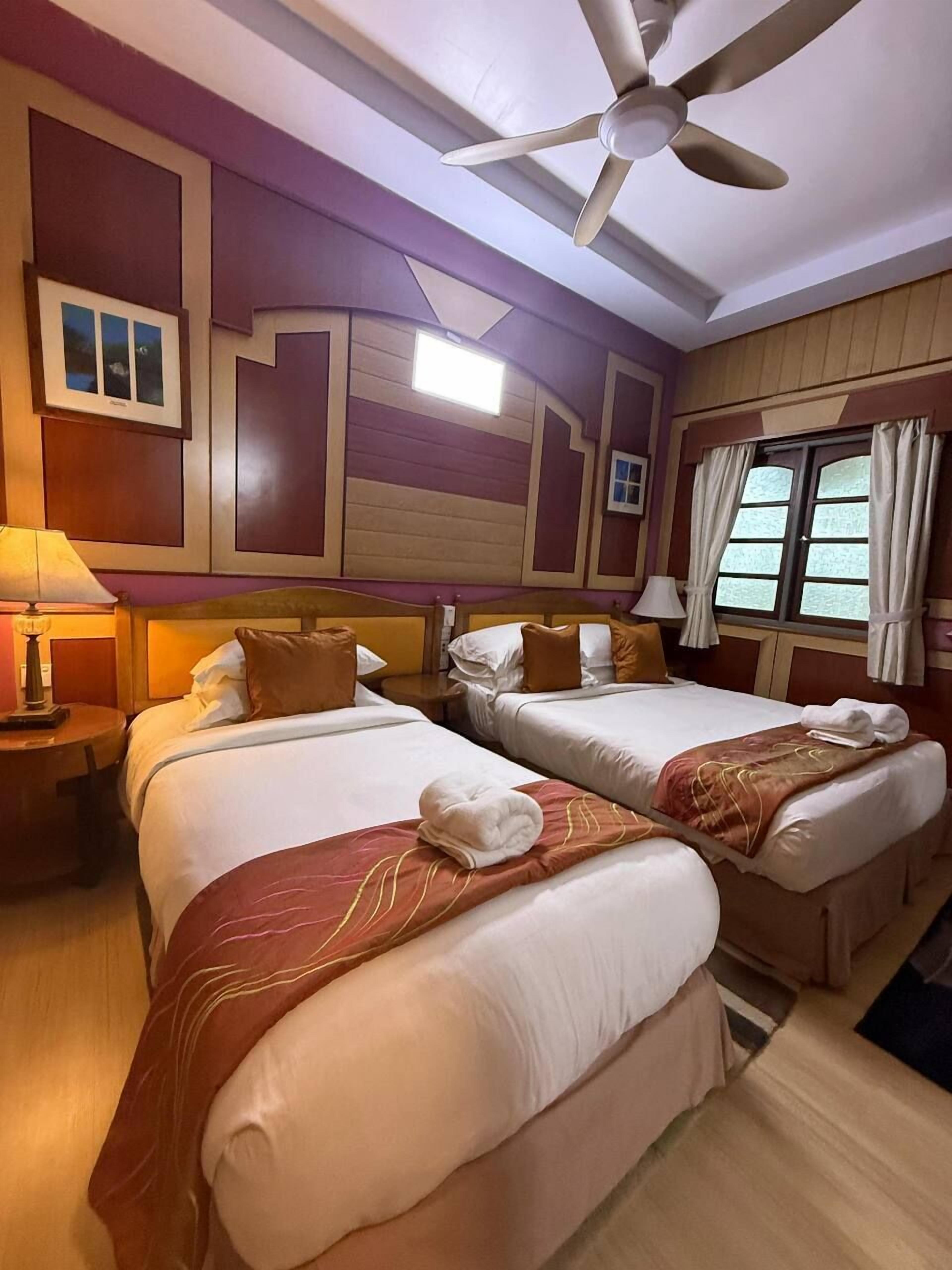 A hotel room with two beds, a ceiling fan, a window with curtains, and a nightstand with a lamp.