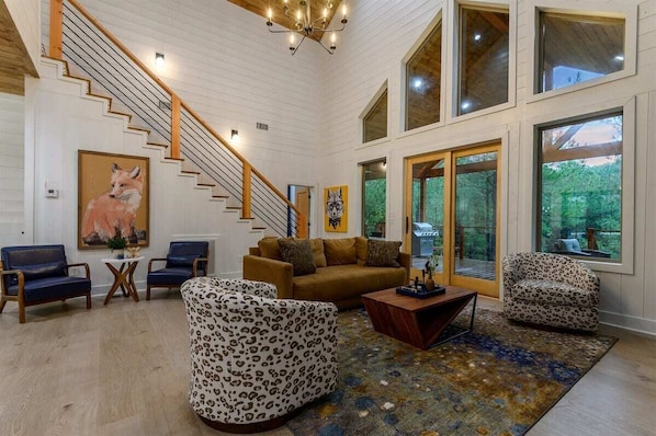 A spacious living room with a staircase, large windows, and a mix of furniture including a sofa, chairs, and a coffee table.