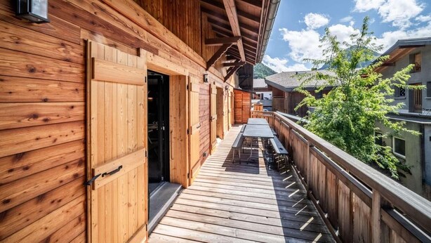 Een houten balkon met een tafel en stoelen, een houten deur en uitzicht op een berg en gebouwen.