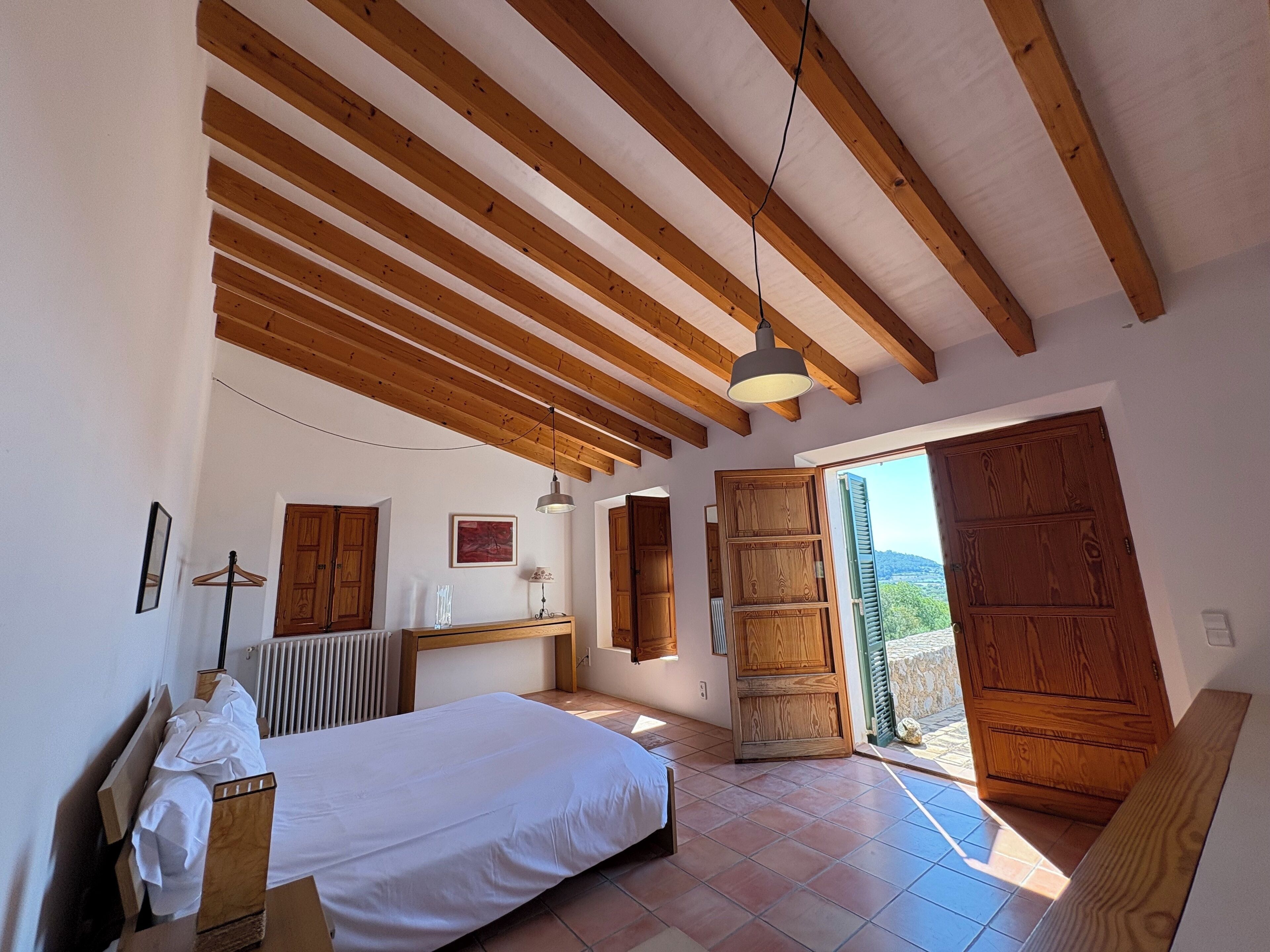 Une chambre avec un lit, des poutres en bois au plafond, un radiateur, un luminaire suspendu et une vue sur l’extérieur par une porte ouverte.