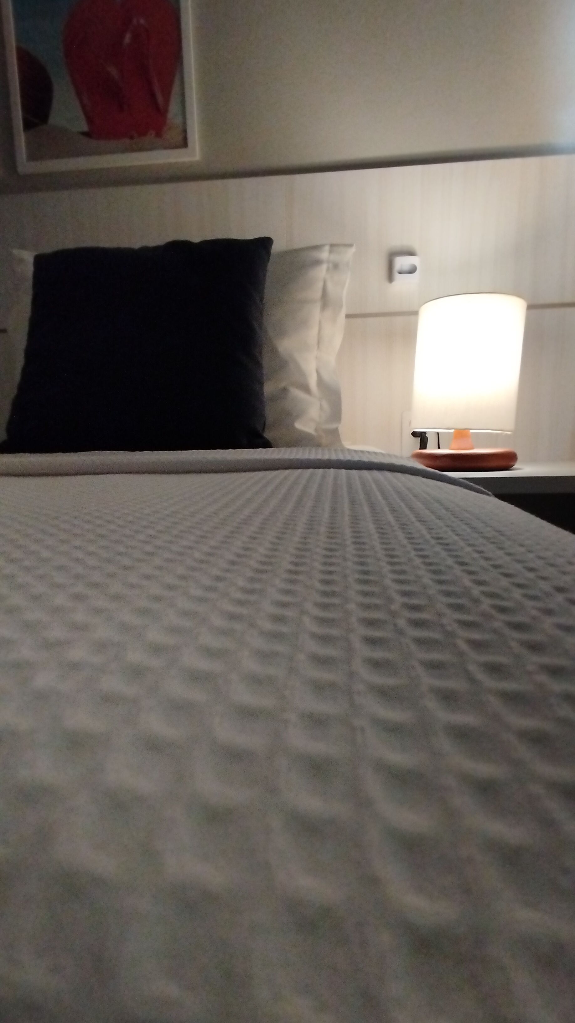 A bed with a textured white cover, a black pillow, a white pillow, and a bedside table with a lamp.