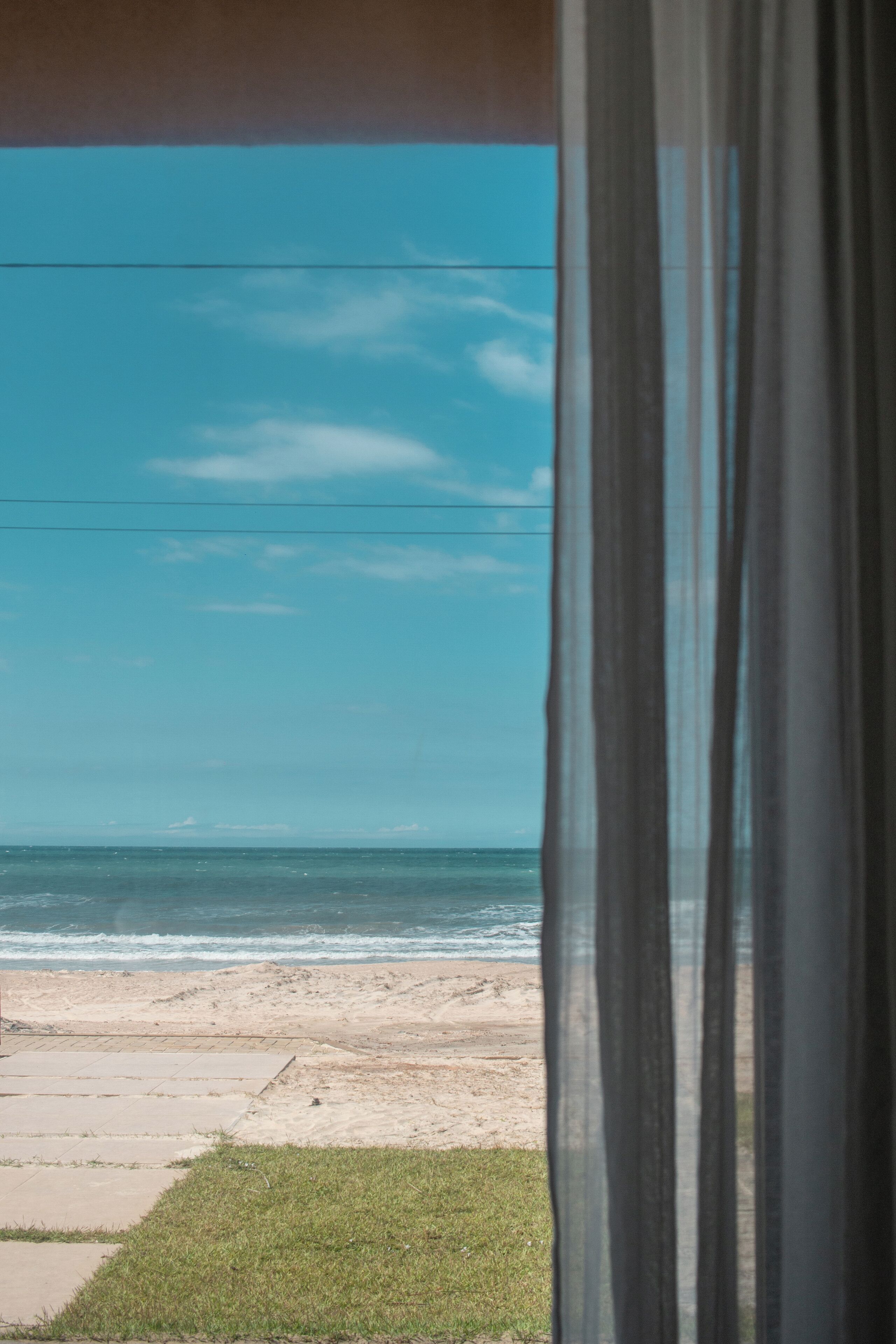 A beach view through a window with curtains, featuring a sandy beach and ocean.