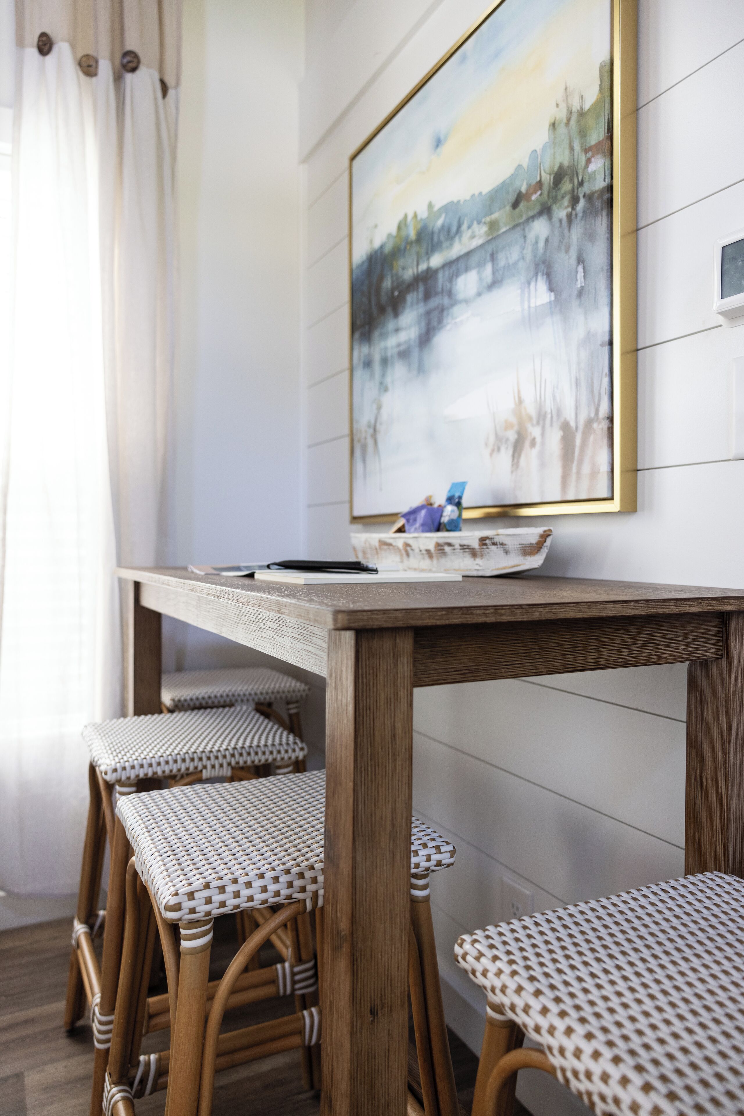 Un coin repas avec une table en bois et des chaises en osier, un tableau au mur et une fenêtre avec des rideaux.