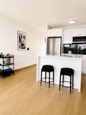 Een moderne keuken met witte kasten, een roestvrijstalen koelkast en een zwart aanrecht. Er staan twee zwarte barkrukken bij en een klein schap met diverse spullen.