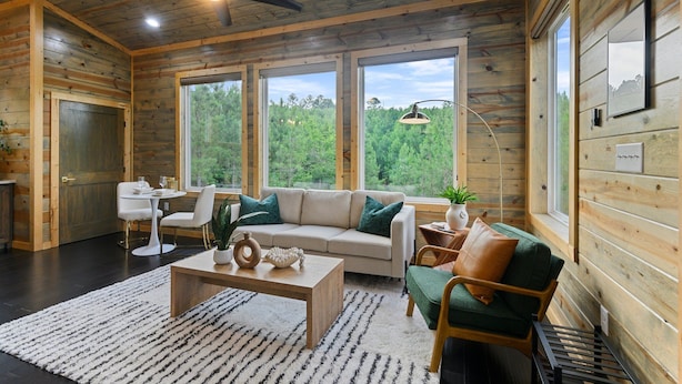 L’intérieur chaleureux d’un chalet avec un plafond en bois, de grandes fenêtres donnant sur la forêt, un canapé, une table à manger et un ventilateur de plafond.