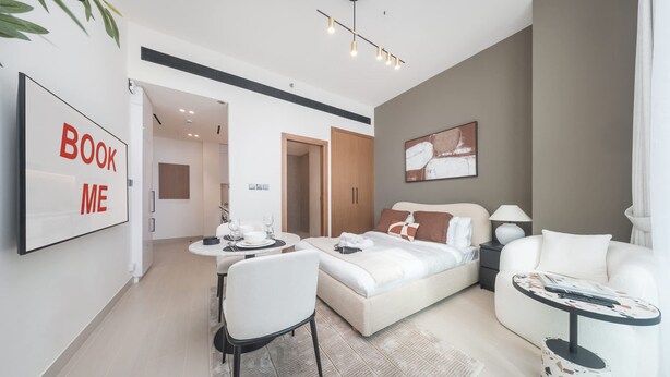 Et moderne hotelværelse med seng, sofa, spisebord og et lille sofabord. Der er et skilt på væggen med teksten 'BOOK ME'.