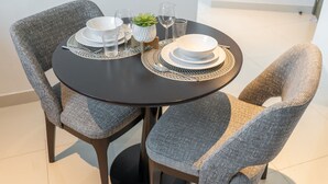 Una mesa de comedor redonda con dos sillas tapizadas en gris, platos blancos, vasos transparentes y una pequeña planta en maceta.