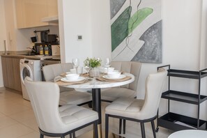Un comedor con una mesa redonda preparada para cuatro personas, rodeada por cuatro sillas. Una estantería metálica negra se encuentra a la derecha.