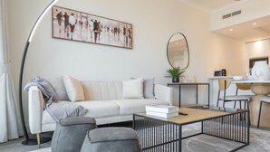 Una sala moderna con sofá, una mesa de centro y sillones grises. Se observa un comedor con mesa y sillas, una cocina al fondo, y un cuadro abstracto de gran tamaño en la pared.