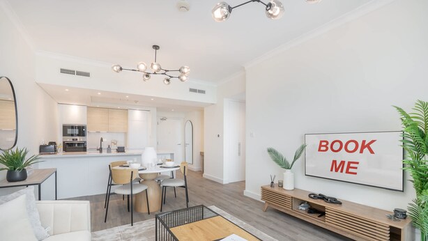 Una sala moderna con sofá, mesa de centro y un comedor con sillas y mesa. Se ve una cocina al fondo con microondas y armarios. Un letrero grande en la pared dice "BOOK ME".
