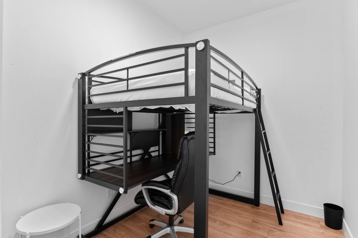 Un lit superposé avec un bureau et une chaise, une petite table ronde et une poubelle, dans une pièce au plancher en bois et aux murs blancs.