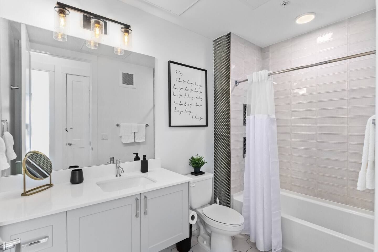 A modern bathroom with a white sink, mirror, and toilet, a shower with a white curtain, and a framed quote on the wall.