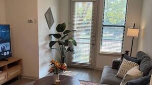 A living room with a gray couch, a coffee table, a TV on a wooden stand, a potted plant, and a window with blinds. - Charming & Cozy 1BR/1BA/2BEDS. Pool/Gym/Shops (Frisco)