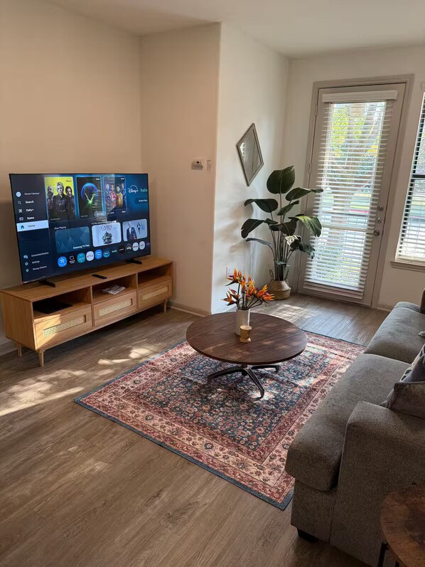 Una sala de estar con un televisor sobre un mueble de madera, una mesa redonda de centro, un sofá, un tapete estampado y una puerta con persianas.