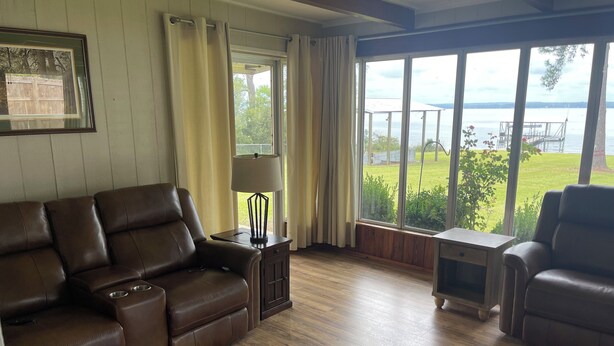 Ein Wohnzimmer mit braunem Ledersofa, einem kleinen Tisch mit Lampe und Blick auf einen See durch große Fenster.