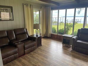 Ein Wohnzimmer mit braunem Ledersofa, einem kleinen Tisch mit Lampe und Blick auf einen See durch große Fenster.