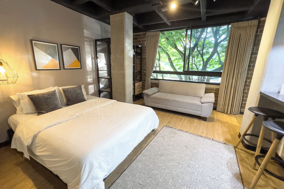 A modern hotel room with a large bed, a sofa, a desk, and a view of trees through the window.