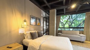 A modern bedroom with a large bed, a nightstand, a sofa, a window with a view of trees, and a hanging light fixture. - Be Together By Roots Rentals (Medellín)