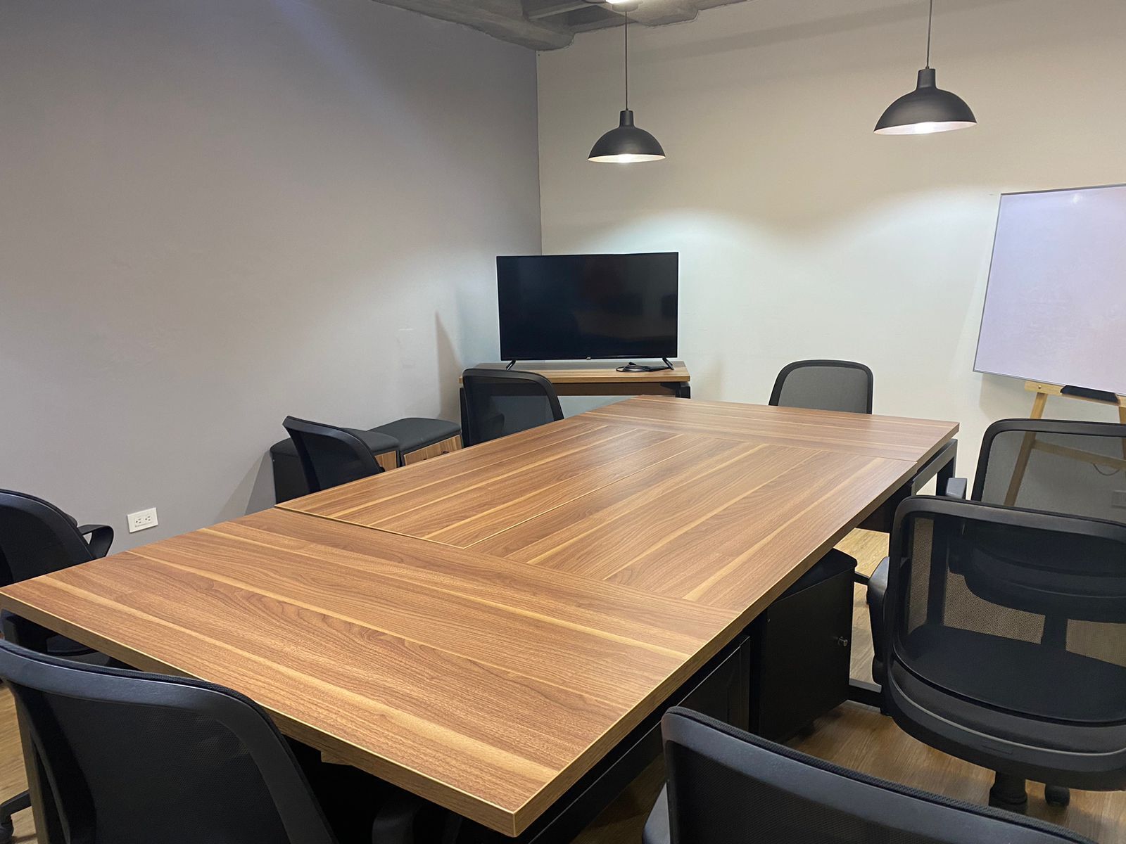 Une salle de conférence avec une longue table en bois, des chaises noires, un téléviseur à écran plat et deux lampes suspendues.