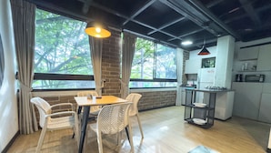 A dining area with a table and chairs, a kitchen area with appliances, and large windows with a view of trees. - Be Together By Roots Rentals (Medellín)