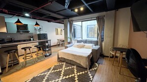 A compact hotel room with a kitchenette, a bed, a dining area, and a large window. - Be Together By Roots Rentals (Medellín)